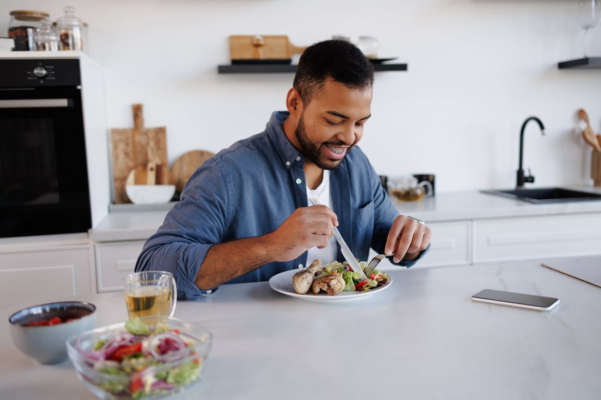 Man smiling while eating a fresh chicken salad with tea and a smartphone in a kitchen