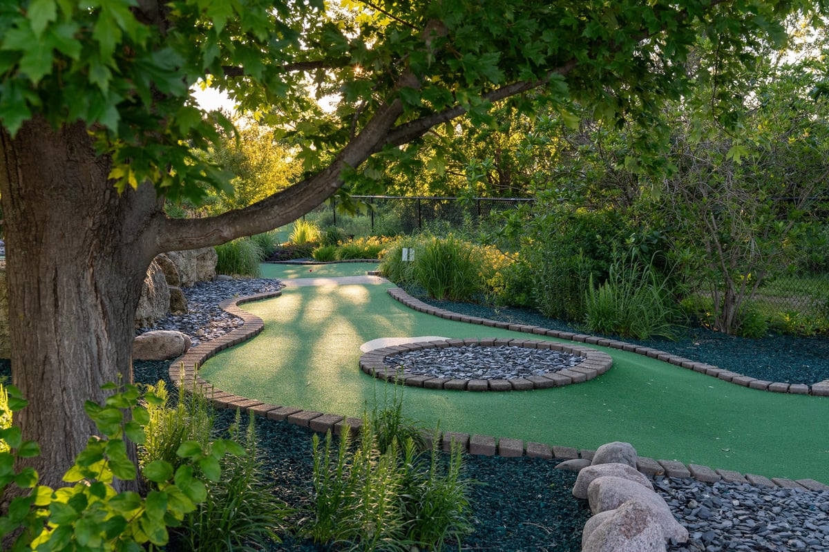 Mini-golf course surrounded by lush greenery, shaded by a large tree