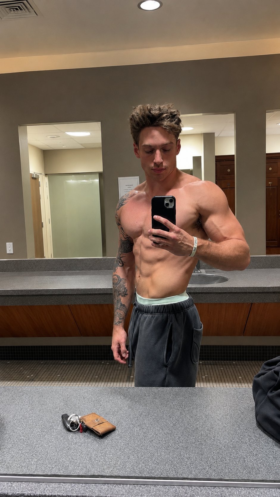 Man taking mirror selfie in locker room, shirtless, holding phone, displaying muscular physique